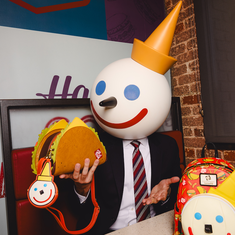 Jack in the Box mascot holding the Loungefly Jack in the Box Taco Crossbody while seated at a table with the Jack in the Box mini backpack to his left.